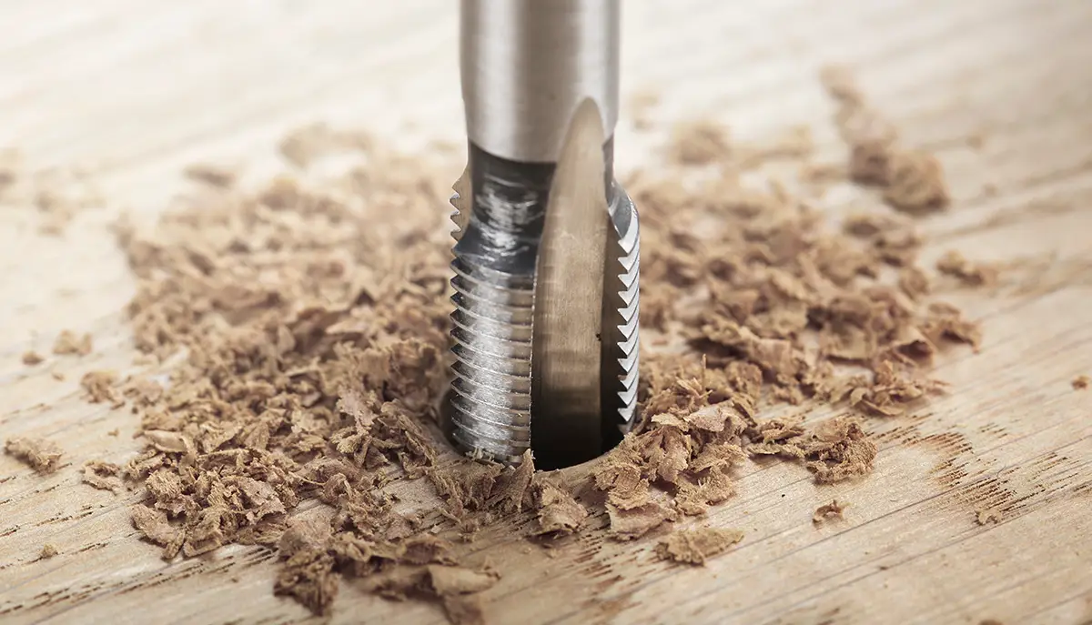 Close-up of a wood thread tap partially inserted into a hole in a wooden board, with wood chips scattered around the borehole.