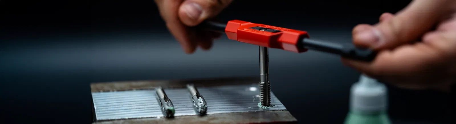Windeisen mit eingespanntem Handgewindebohrer, womit ein Gewinde in einen Metallblock geschnitten wird. Daneben liegen zwei weitere Handgewindebohrer.