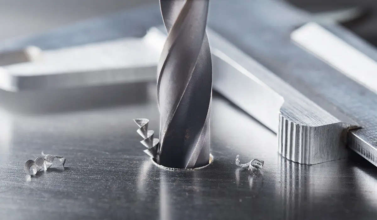 Spiralbohrer bohrt Loch in Stahlplatte. Späne und Messschieber liegen daneben.